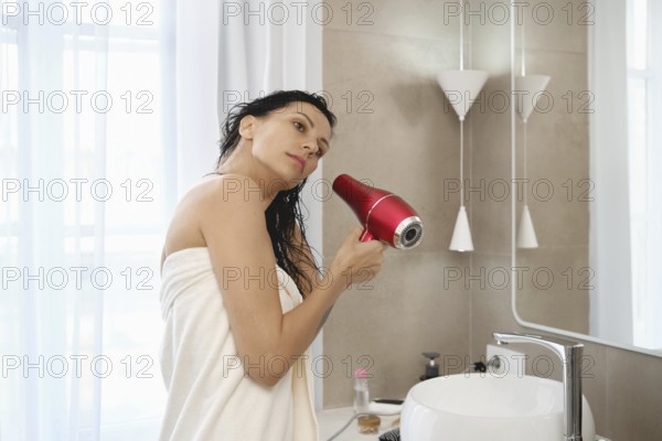 A woman stands in a well-lit bathroom, wrapped in a towel and using a vibrant red hair dryer to dry
