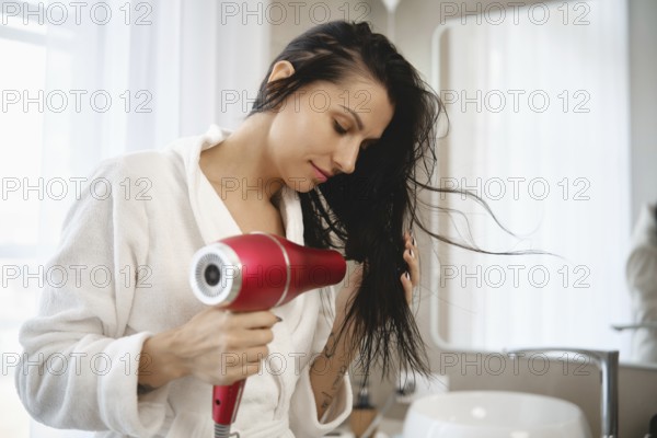 A woman wearing a white bathrobe is focused on drying her long, wet hair with a red hairdryer. The