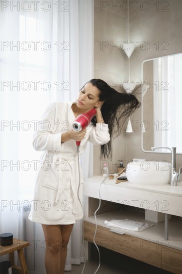 A woman stands in a modern bathroom, using a hair dryer to style her long, wet hair. She is dressed
