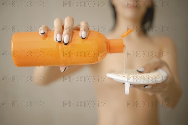 A person with wet hair uses an orange bottle to dispense liquid soap onto a textured sponge. The