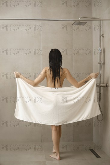 A woman stands in a shower holding a large white towel. Water droplets linger on her wet hair. The