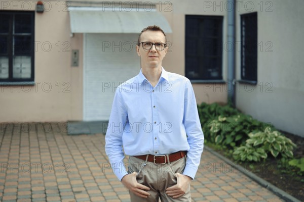 A man with glasses in a light blue shirt and beige trousers standing in front of a building. He has