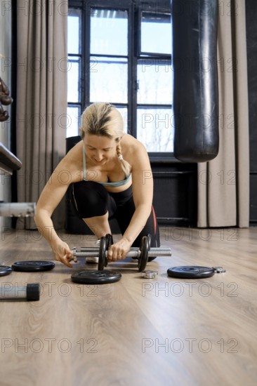 A focused woman is adjusting weights while kneeling on a gym floor. The space has large windows