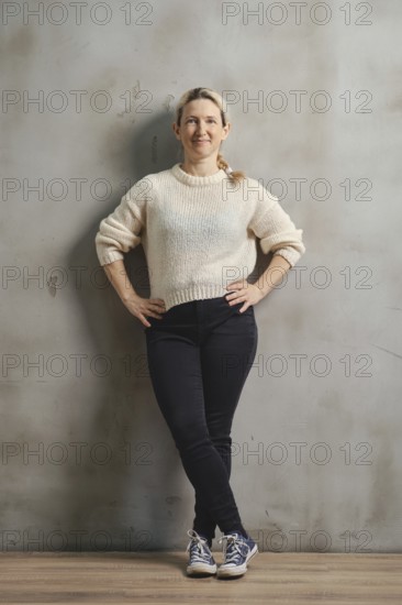 A woman stands casually with her hands on her hips, wearing a soft sweater and dark jeans. She