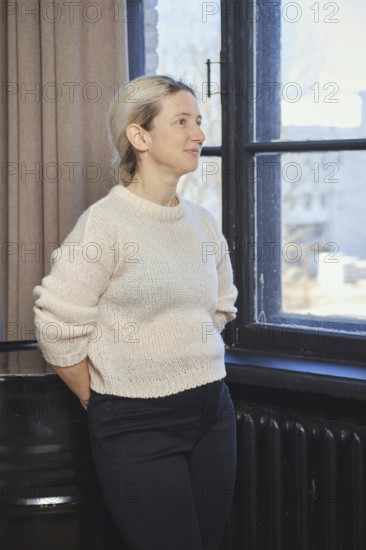 A woman with light hair is standing by a window, gazing thoughtfully outside. She is dressed in a