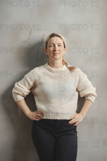 A woman with long, light hair stands upright with her hands on her hips, wearing a soft beige