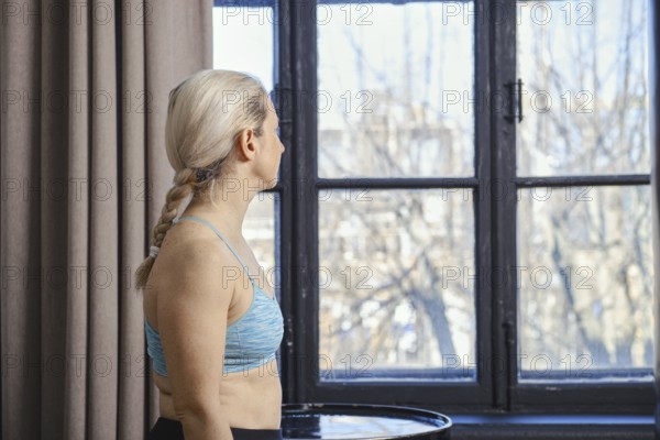 A woman with braided hair is standing near a large window, looking outside at the bright day. She