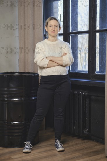 A woman stands with arms crossed, dressed in a white sweater and dark pants, exuding confidence in