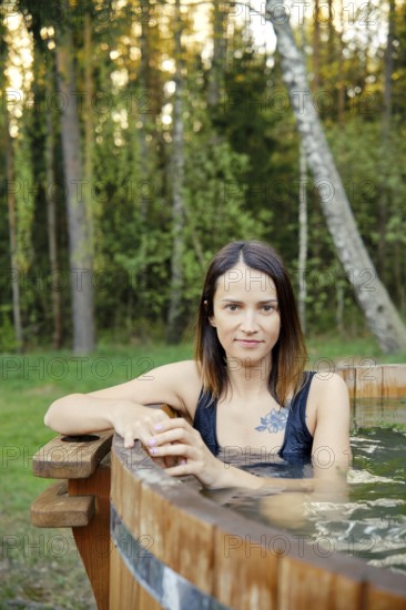 Outdoor portrait of pretty woman in a wooden hot tub surrounded by lush greenery. The warm glow of