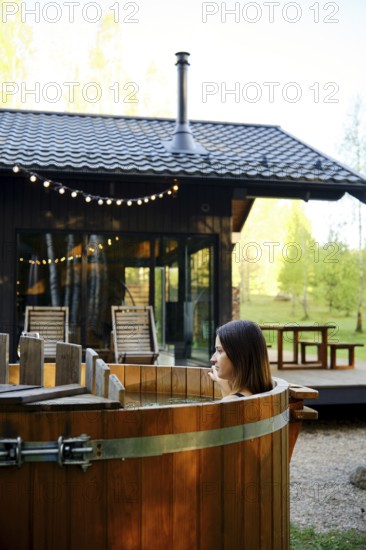 A woman enjoys relaxing in a wooden hot tub in the backyard of a tiny house. The sun illuminates