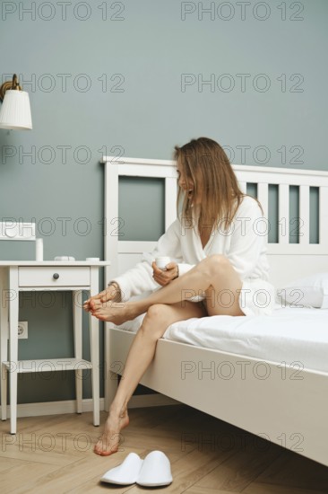 A woman in a white robe smiles while applying lotion to her feet in a charming bedroom. Natural