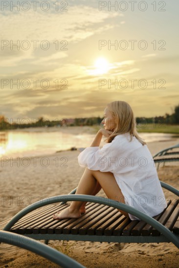 A woman sits pensively on a wooden lounge chair by the lakeside as the sun sets. She gazes at the