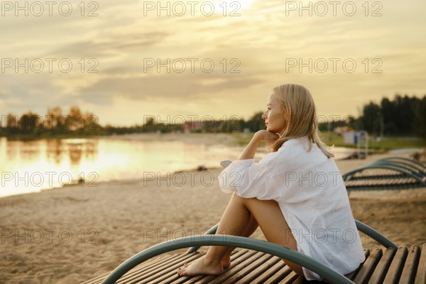 A woman sits on a wooden bench by the shore, gazing at a stunning sunset over the water. The soft
