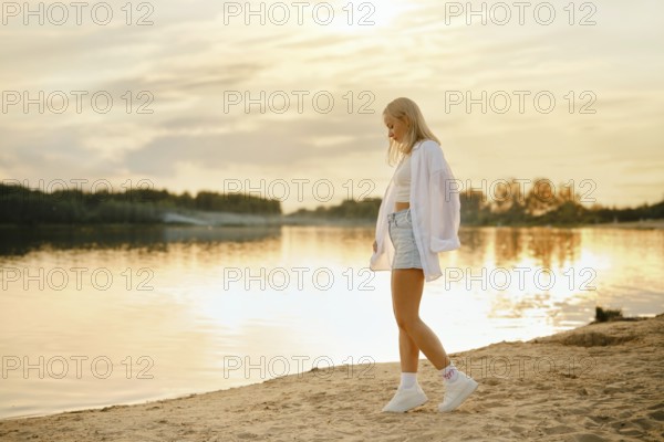 A young woman strolls on the sandy beach at sunset, with the golden light reflecting on the calm