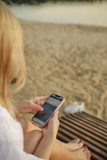 A woman sits on a wooden lounge, absorbed in her smartphone. The sun sets in the background,