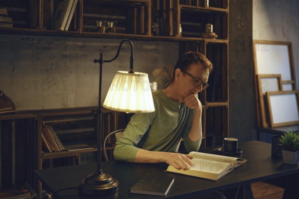 A man sits at a desk in a dimly lit room, engrossed in a book. Warm light from a lamp highlights