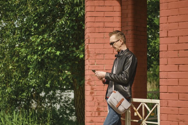 A man wearing sunglasses and a leather jacket stands by red brick columns, attentively using his