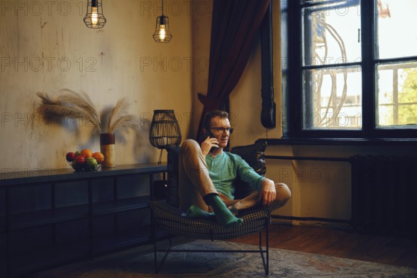 A man dressed in a green long-sleeve shirt and light pants relaxes in a chair, engaging in a phone