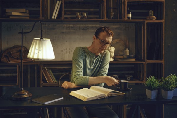 A man enjoys a quiet evening in a rustic home office, reading a book while sipping herbal tea. The
