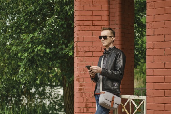 A man dressed in a leather jacket and sunglasses stands by red brick columns. He holds a smartphone
