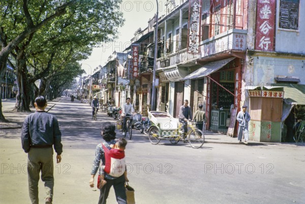 People walling and cycles in tree lined street with small shops, Macau, Asia 1964