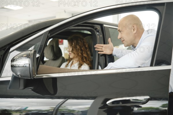 A woman evaluates a car while seated inside it at a car dealership. A salesperson gestures towards