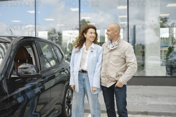 A couple stands beside a sleek black car at a bustling car dealership, engaged in a lively