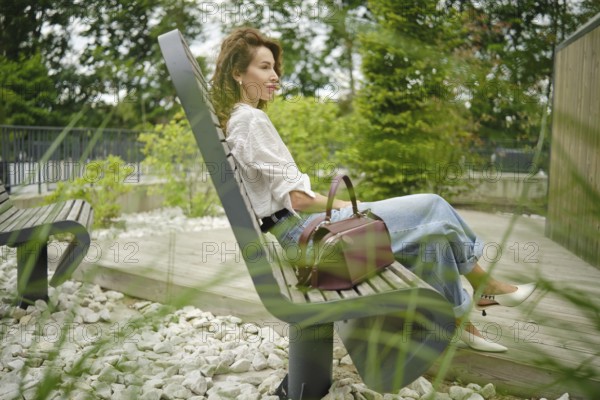 A woman sits comfortably on a bench in a park, surrounded by greenery and decorative stones. She