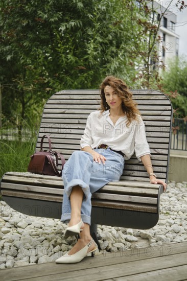 A woman sits comfortably on a unique wooden bench in a modern urban garden. She wears casual