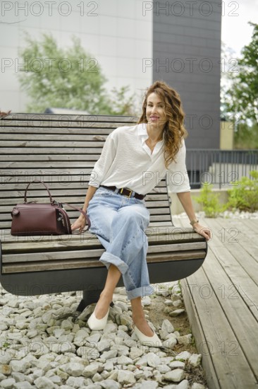 A woman sits comfortably on a modern bench in a lush park, wearing a light button-up shirt and