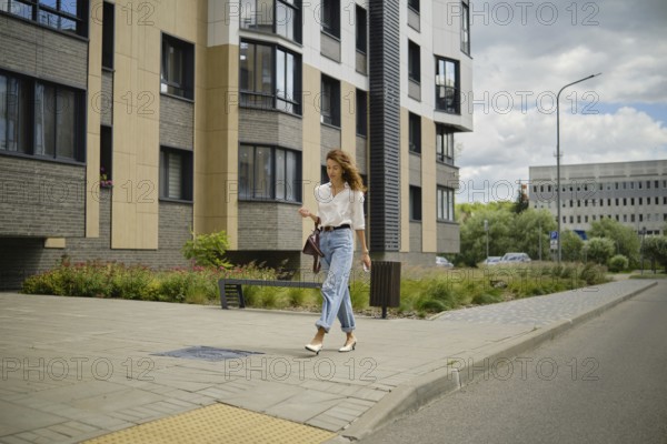 A woman strolls down a city sidewalk on a sunny day. She wears a casual outfit with light jeans and