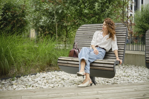 A woman with curly hair is seated on a modern wooden bench in a landscaped urban park. She is