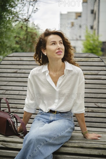 A woman with curly hair sits relaxed on a wooden bench, wearing a white shirt and blue jeans. She