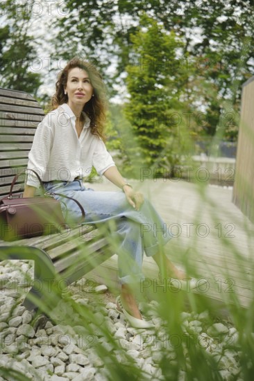 A woman sits calmly on a wooden garden bench, dressed in a light shirt and jeans. Tall grass sways