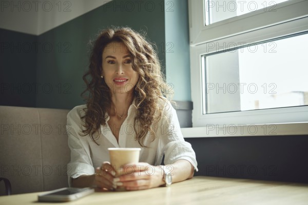 A woman with curly hair sits at a wooden table in a cafe, holding a cup of coffee. She smiles while