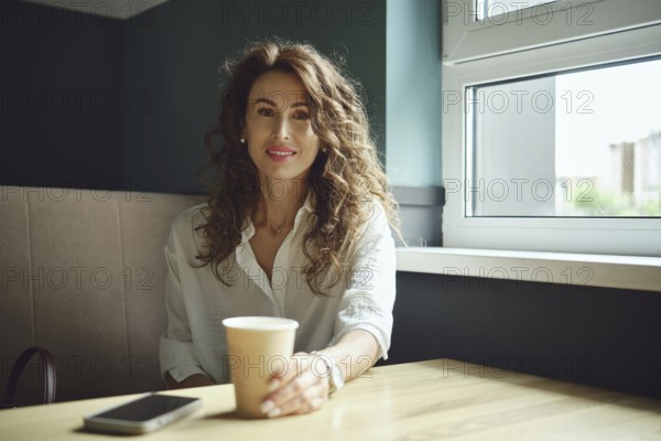 A woman with curly hair sits at a table near a window, holding a cup of coffee. She wears a white