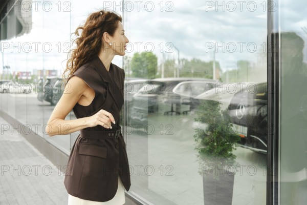 A woman stands beside a large glass window in a car showroom, gazing at displayed vehicles. Her