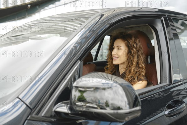 A woman with curly hair is smiling as she sits in the driver's seat of a sleek car. The vehicle is
