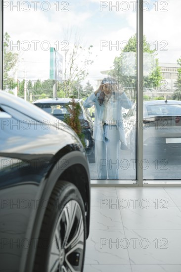 A woman stands outside looking in through large glass windows to a car dealership, admiring various