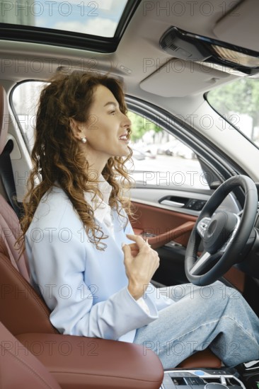 A woman with curly hair is seated in the driver's seat of a modern car, smiling and looking forward