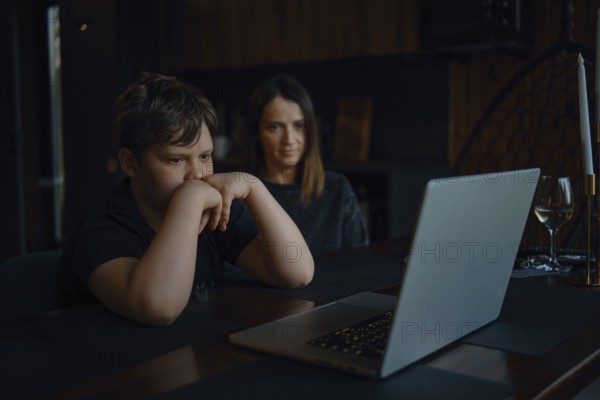 A boy sits focused at a table, watching a laptop screen closely while his mother looks on with a