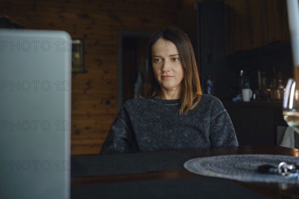 A woman is focused on her laptop while sitting at a wooden table in a warm dark living room. The