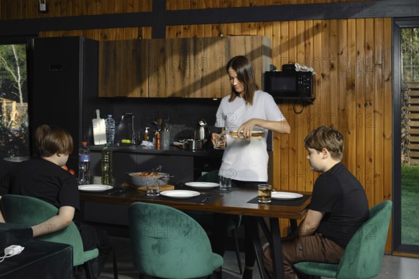 In the kitchen, a woman pours a drink, while two children are sitting at the dining table,