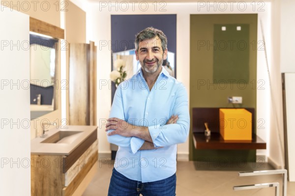 Confident salesman smiling with crossed arms while posing in a bathroom furniture store, showcasing