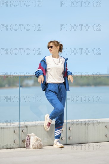 A woman relaxes by a glass railing at the waterfront on a bright sunny day. She wears a colorful