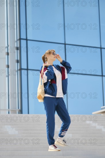 A young individual stands on a staircase, wearing sporty clothing and sunglasses. The sunny weather