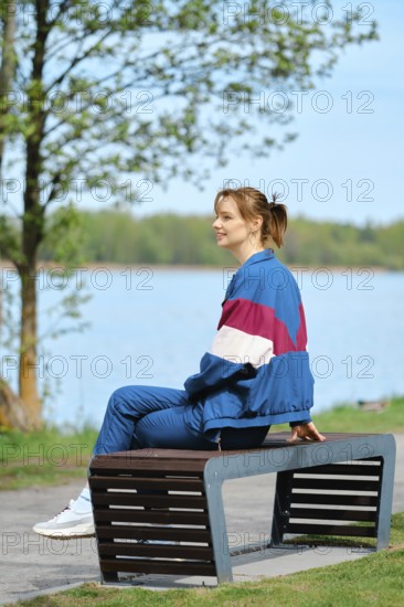 A young woman relaxes on a modern bench near a serene lake, dressed in colorful activewear. The