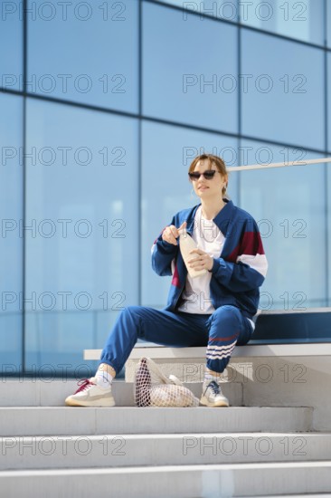 A young woman dresses in a stylish tracksuit sits on outdoor steps near a contemporary structure.