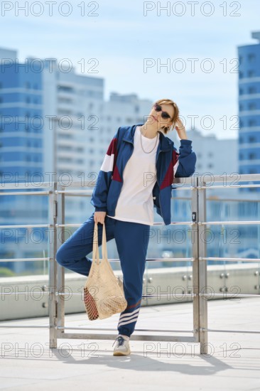 A woman dressed in a sporty outfit stands confidently on a terrace surrounded by a bright blue sky
