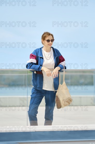 A woman stands by a glass railing at a waterfront location, dressed in a colorful jacket and